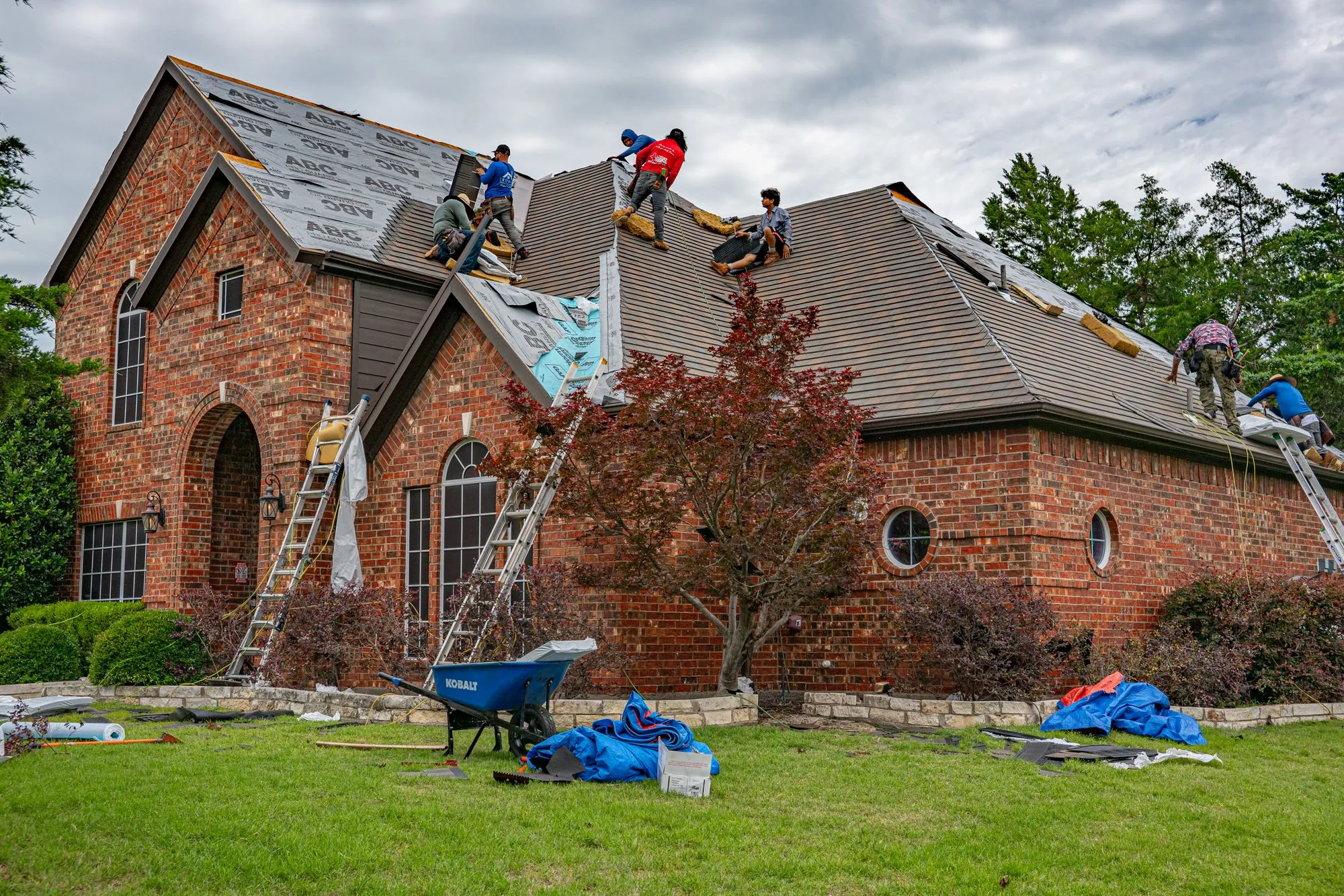 A crew working across the slopes of a brick house, wheelbarrow and tarps out on the lawn