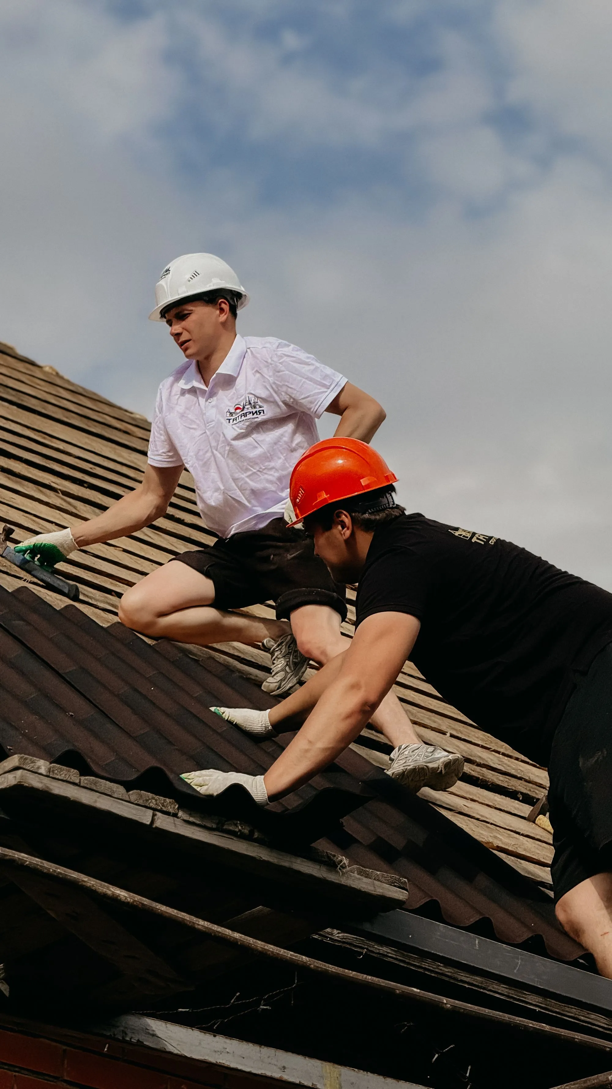 Two roofers in hard hats stripping a course of shingles together