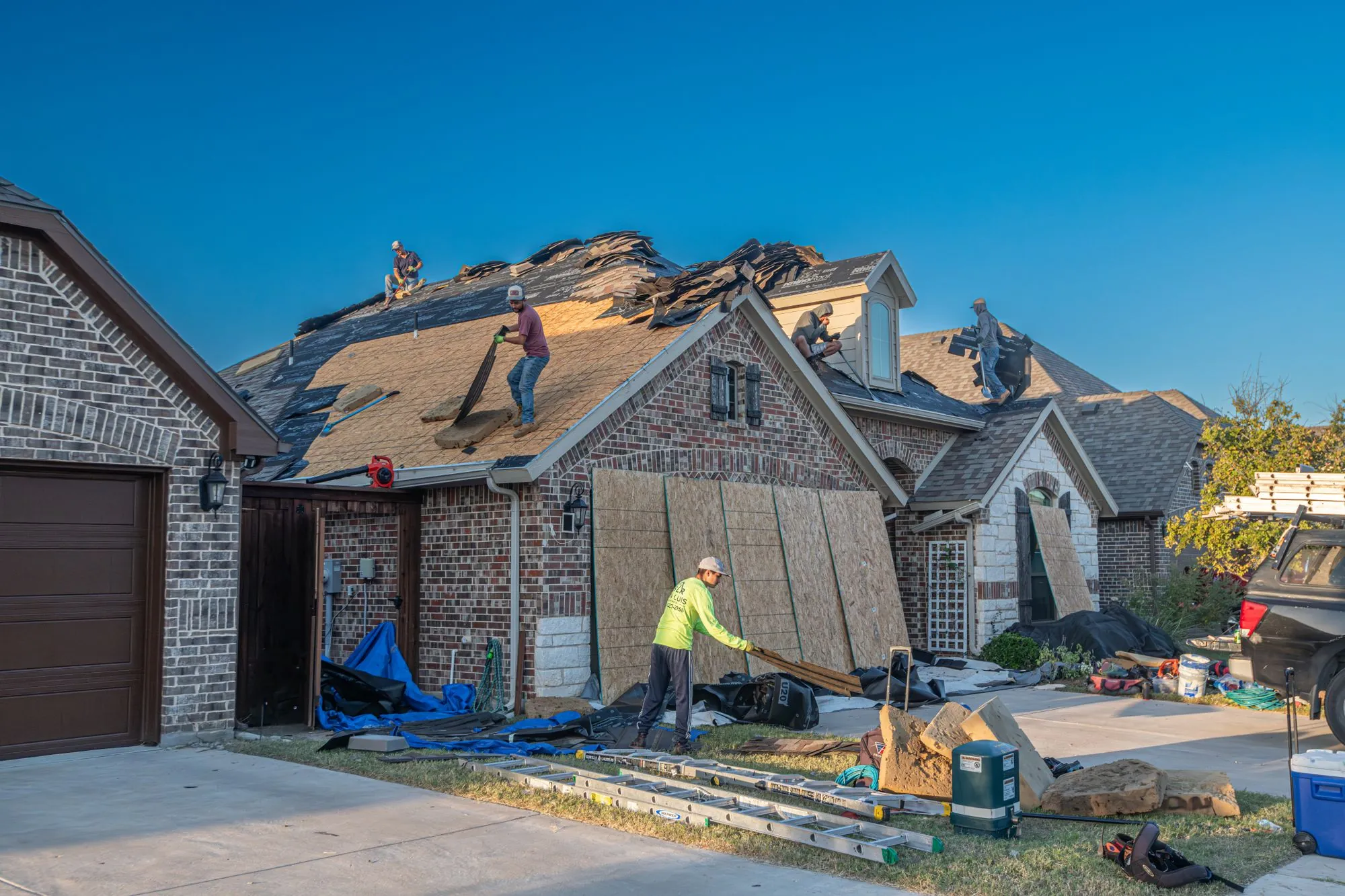 A roofing crew working across the slopes of a large brick house, ladders up and the drive sheeted over