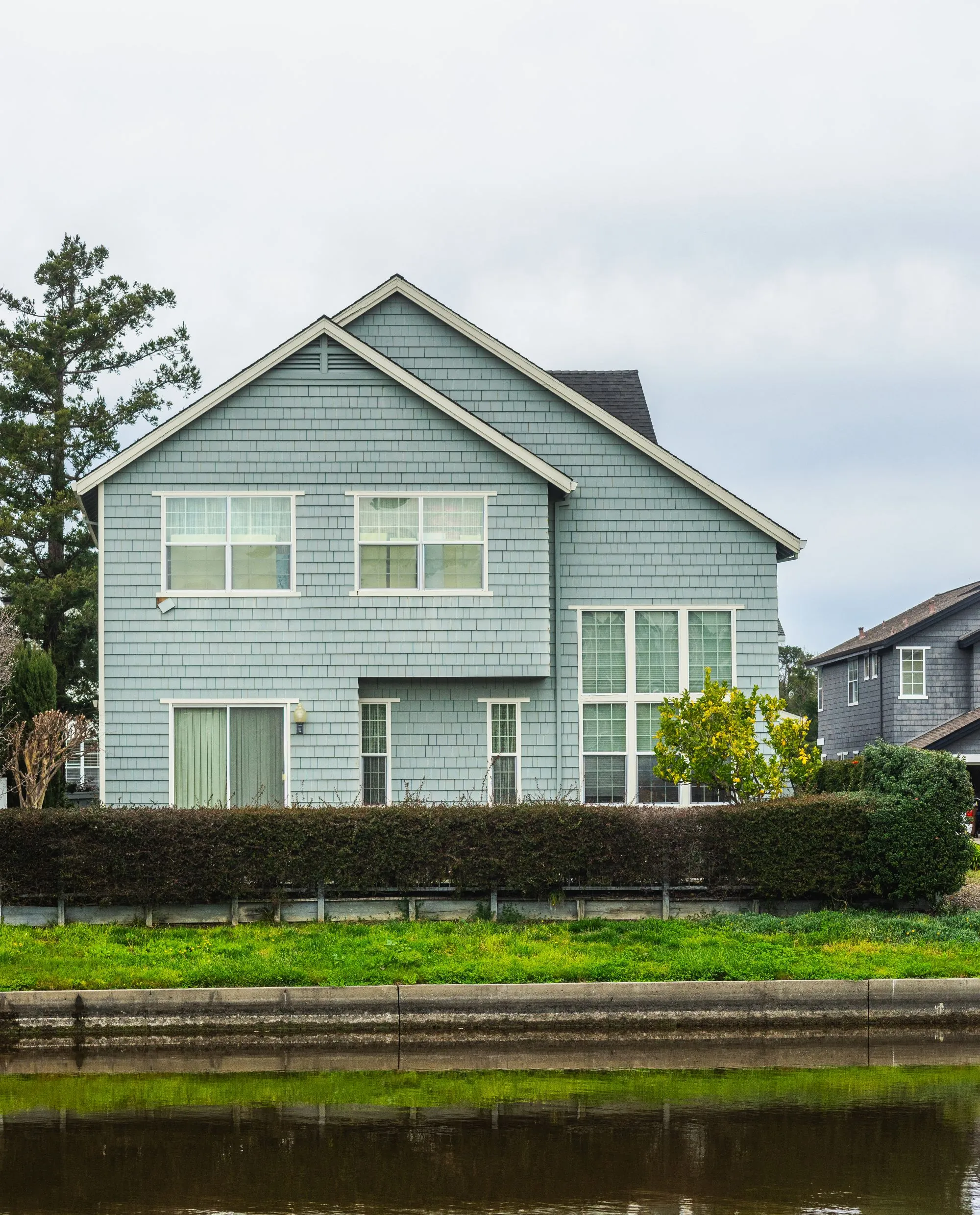 A pale blue two-storey house with a shingled roof beside still water